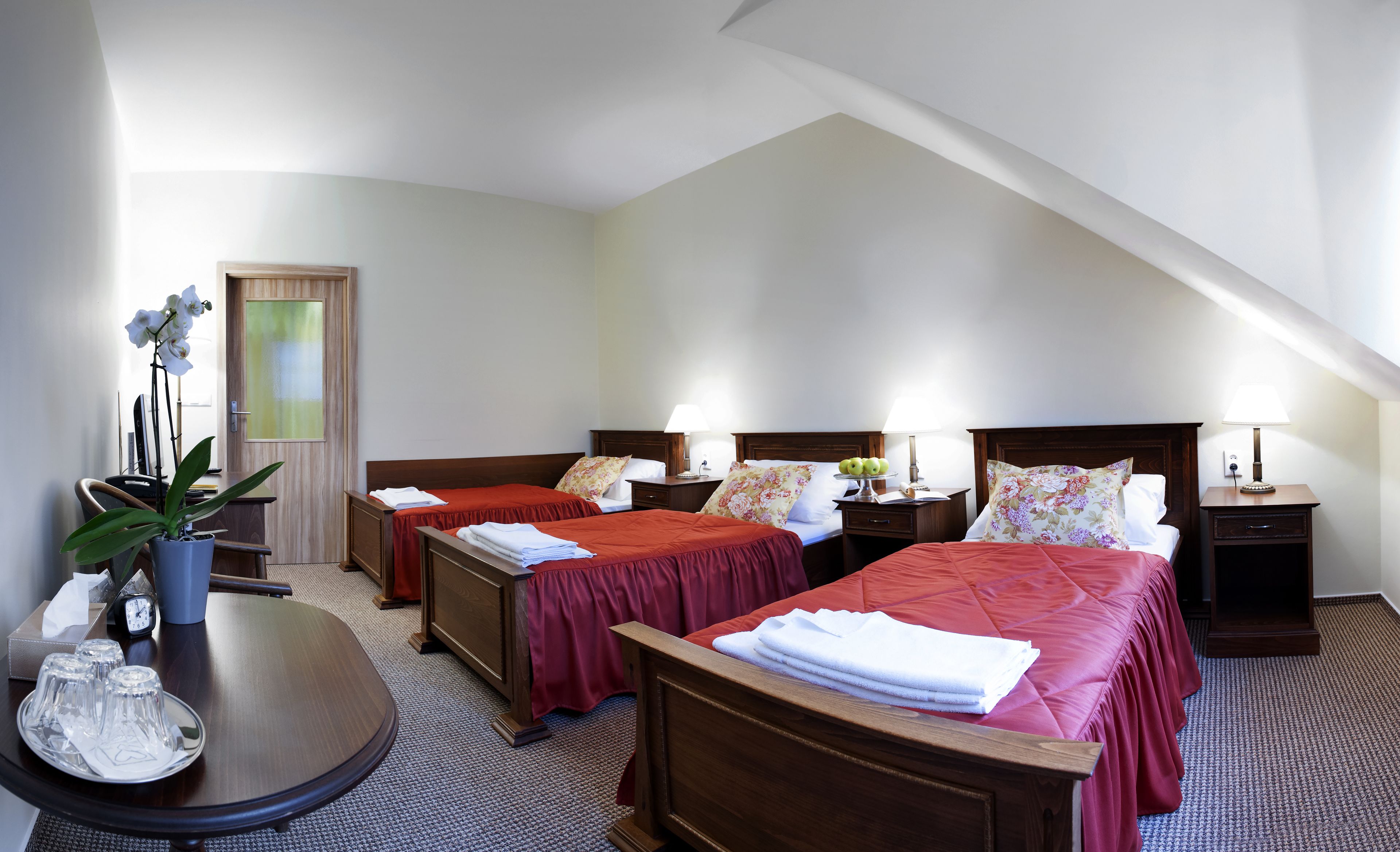 A hotel room with two single beds, a wooden desk, a chair, a small table with a plant, and a clock.