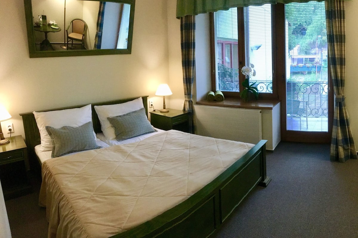 Ein Hotelzimmer mit Bett, Schreibtisch, Stuhl, Lampe, einem Fenster mit Vorhängen und einem Bild an der Wand.