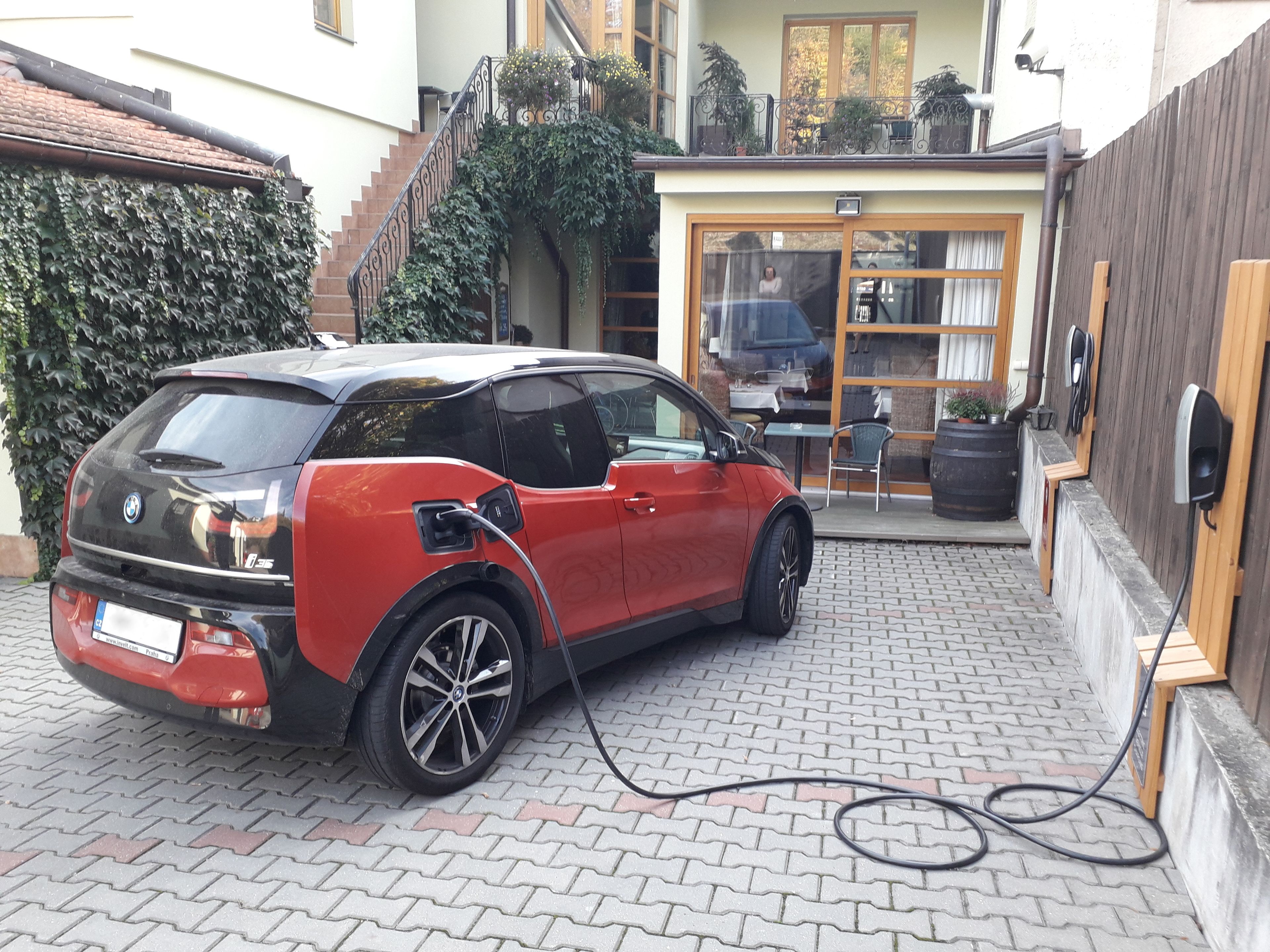 A red BMW i3 charging in a courtyard with a brick floor, a wooden fence, and a building with a glass door in the background.
