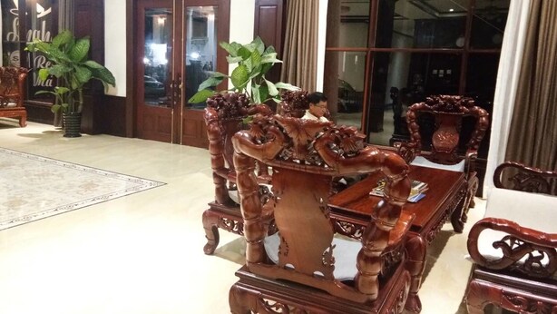 A traditional wooden dining set with intricate carvings, a large potted plant, and a view of the outside through a glass door. - Singha Hotel (Oudomxay)