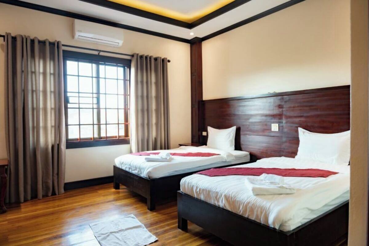 Ein Hotelzimmer mit zwei Betten, Holzboden, einem großen Fenster mit Vorhängen und einer Klimaanlage.