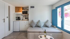 A compact kitchen with a microwave, coffee maker, and hanging utensils. A small seating area with cushions and a table with wine glasses and a bottle is visible. - Euphoria Suites (Santorini)