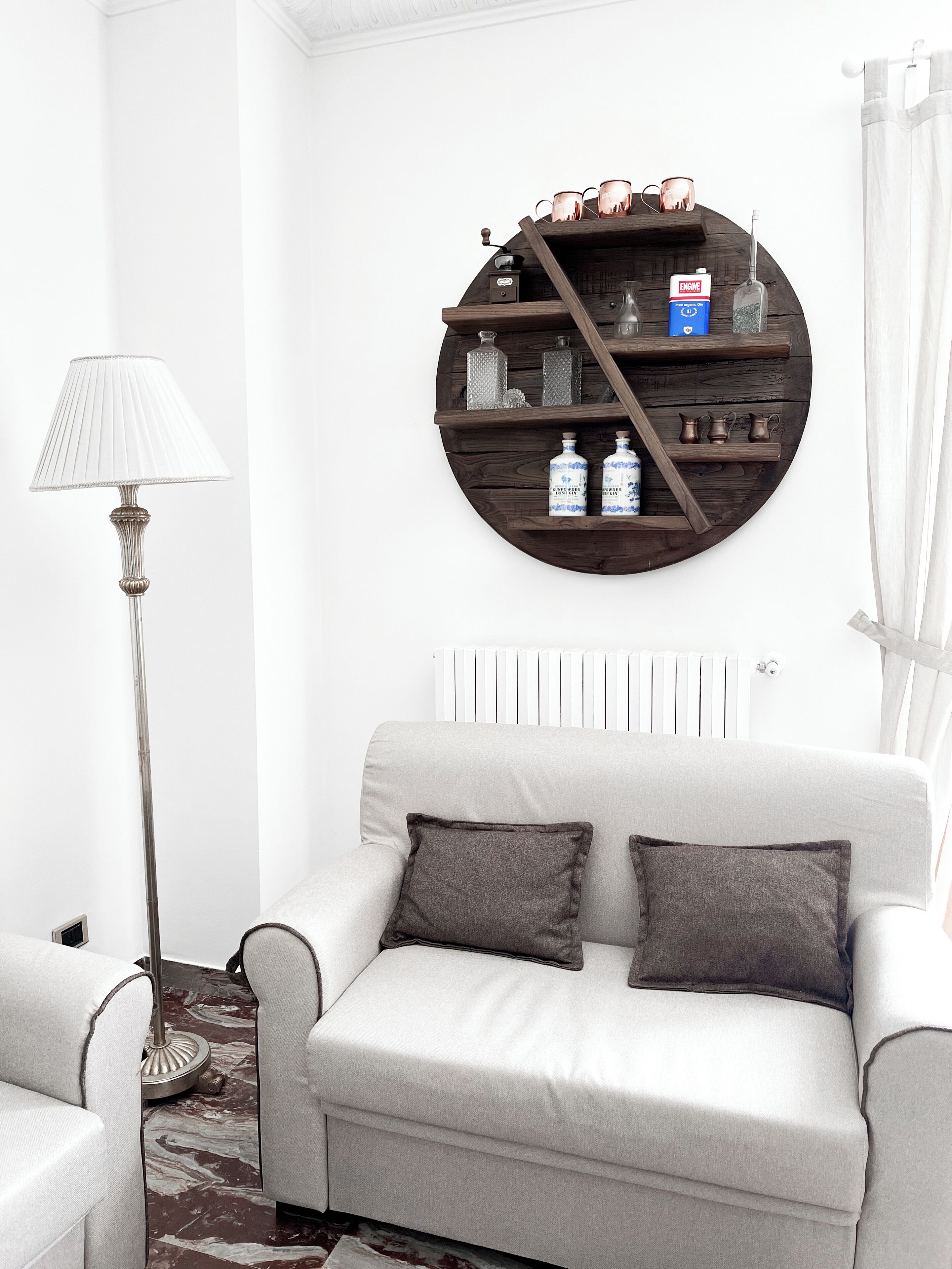 A modern living room with a round wooden shelf displaying various items, a white sofa with two cushions, and a floor lamp.