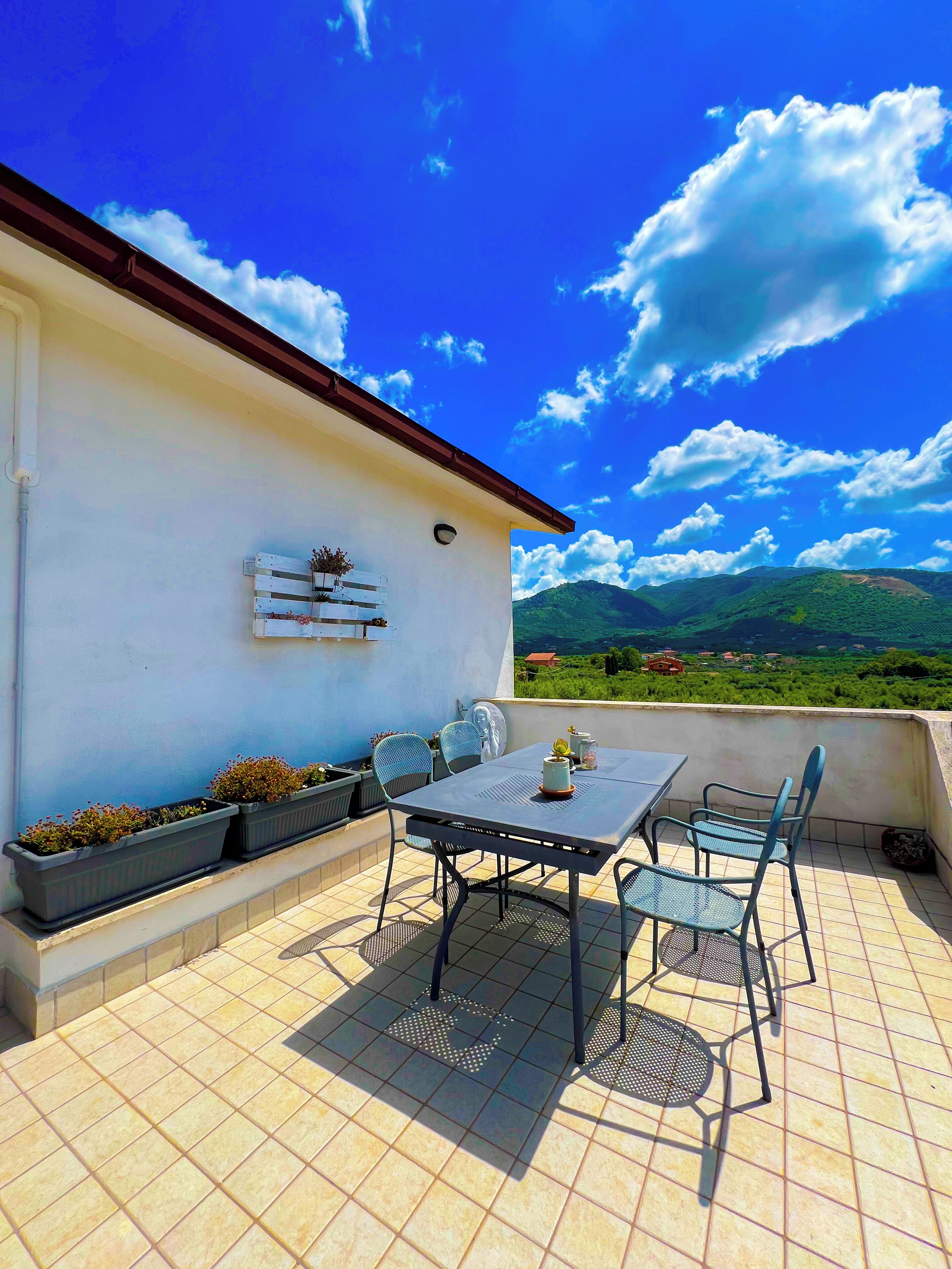 An outdoor patio with a table and chairs, potted plants, and a view of green hills.