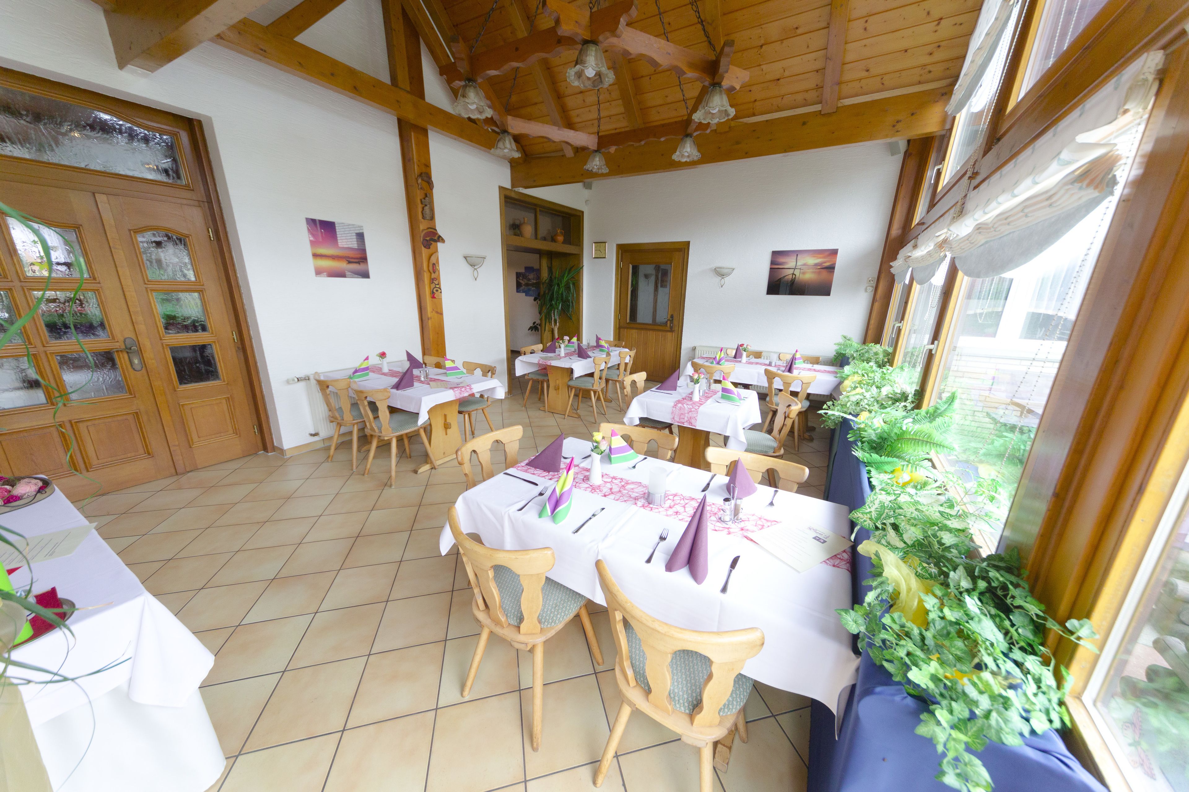 A dining area with wooden tables and chairs, a tiled floor, and a view of the outdoors through large windows.