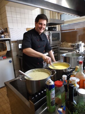 Ein Koch in einer Küche mit Edelstahlgeräten, einer Mikrowelle sowie verschiedenen Kochzutaten und Utensilien.