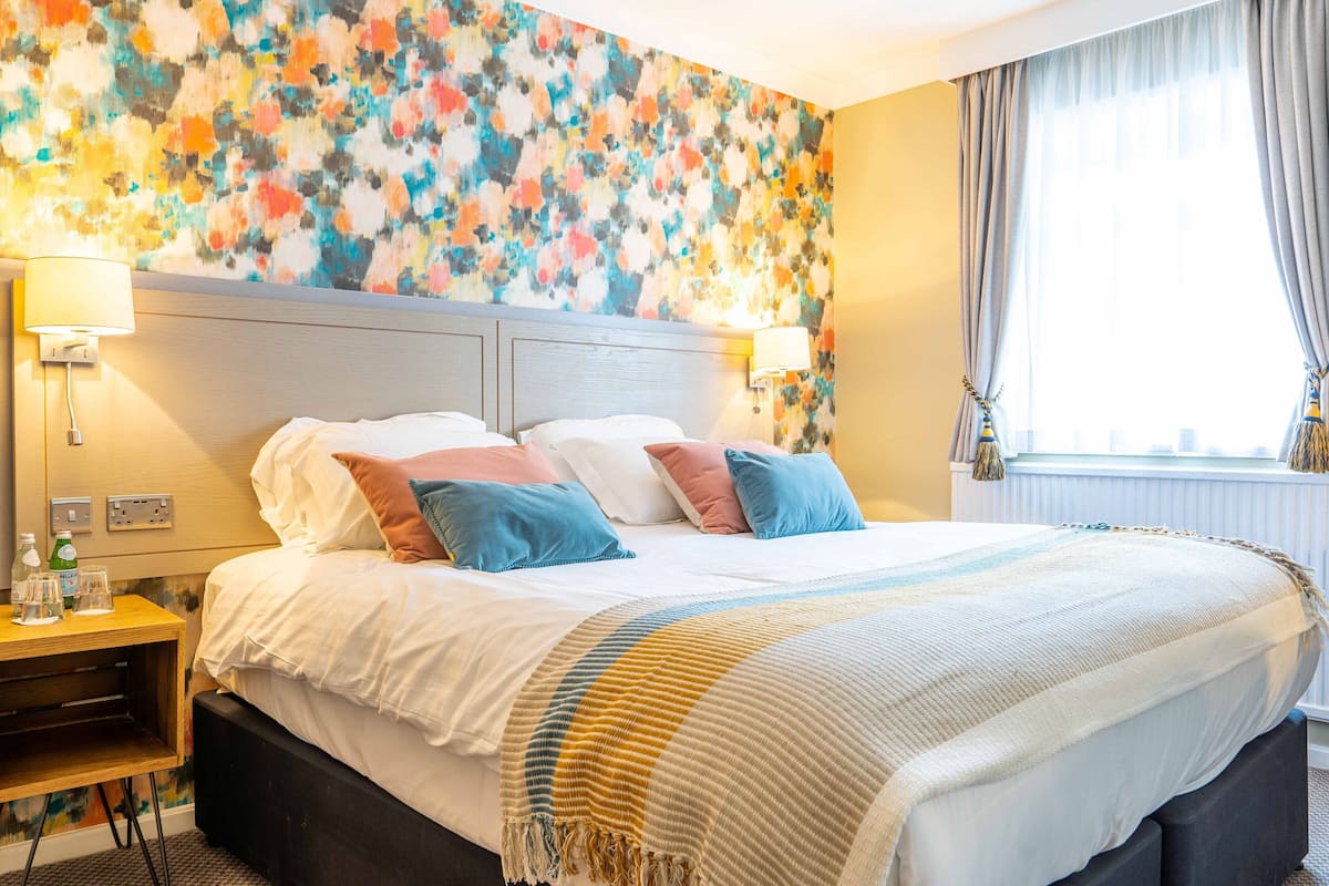 Una habitació d'hotel amb un llit gran, una tauleta de nit, una làmpada i una finestra amb cortines.