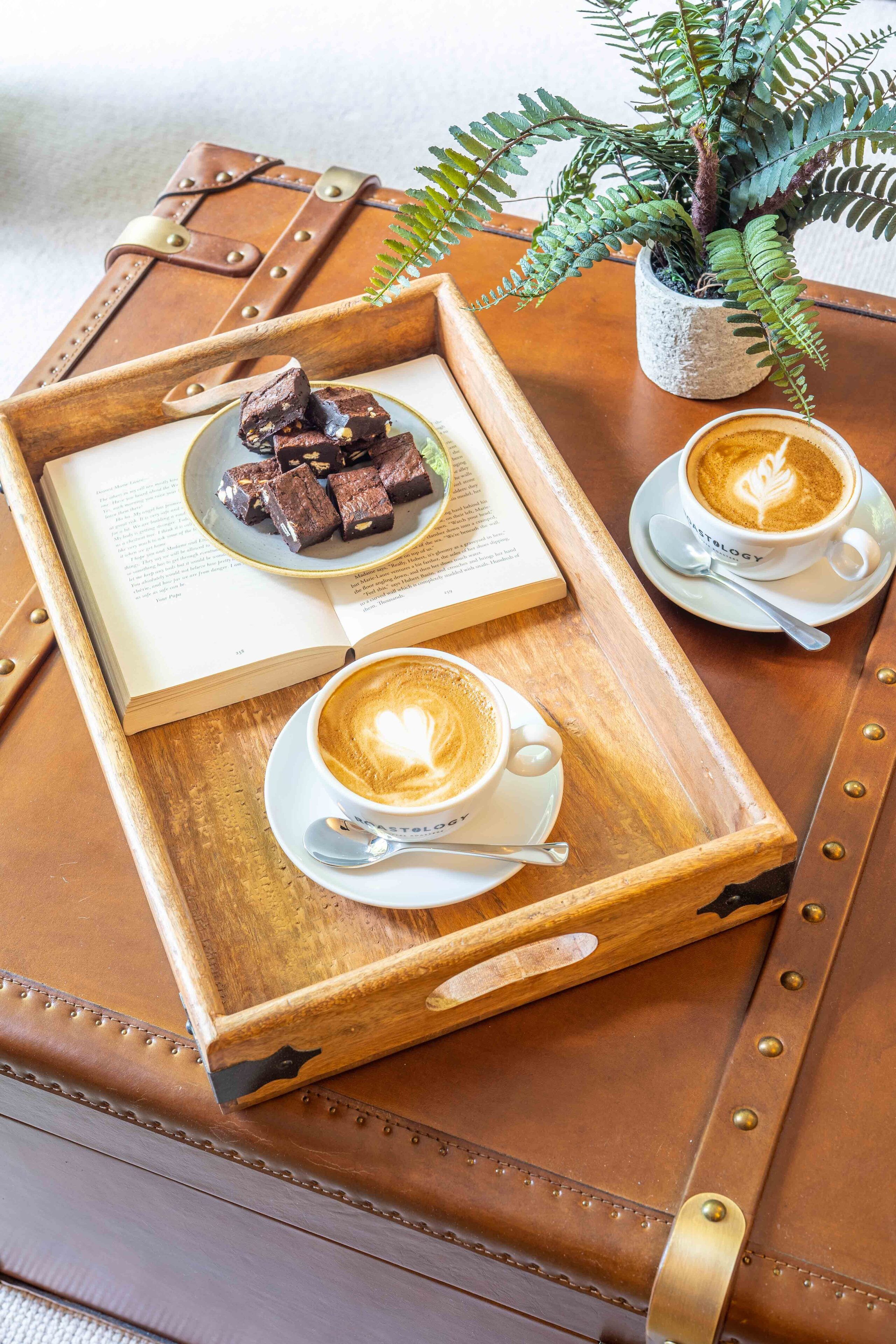 A wooden tray with a book, two cups of coffee, and a plate of brownies on a leather-bound suitcase.