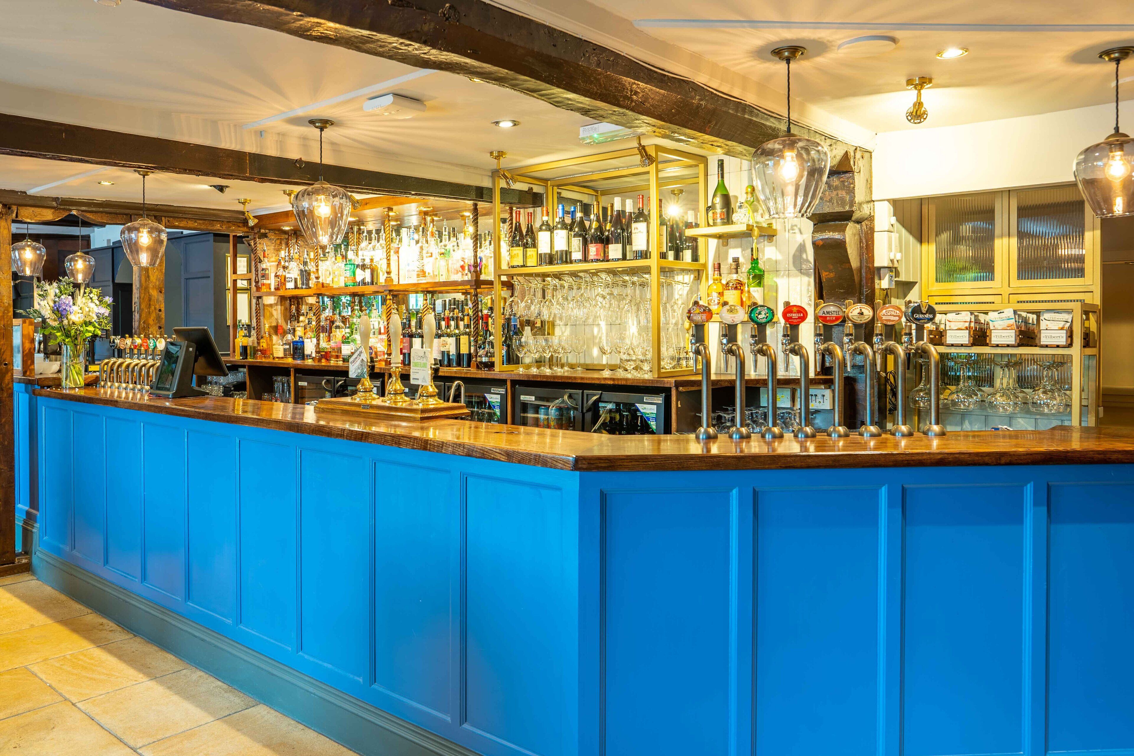 A bar area with a blue counter, various bottles, glasses, and a selection of beers on tap.