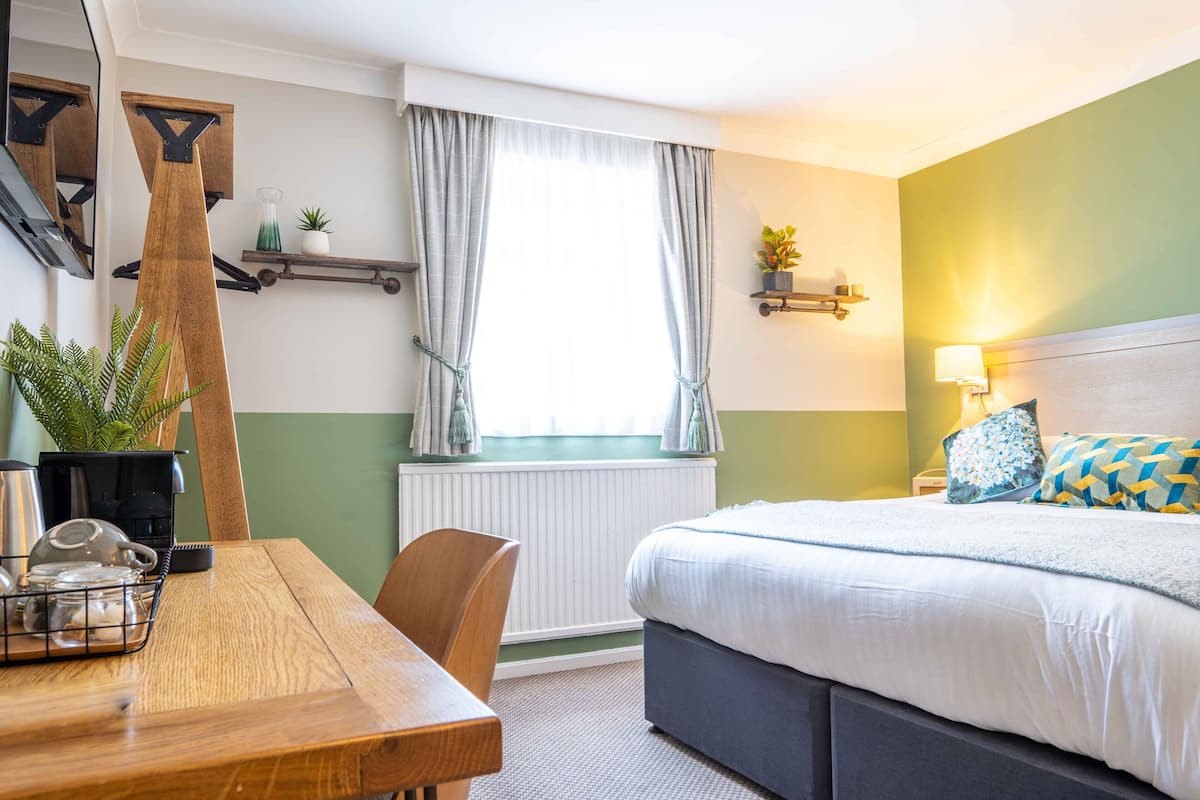 Habitació d'hotel amb un llit, una taula de fusta amb una tetera, una cadira, un radiador, una finestra amb cortines, una estant amb plantes i una làmpada penjada a la paret.