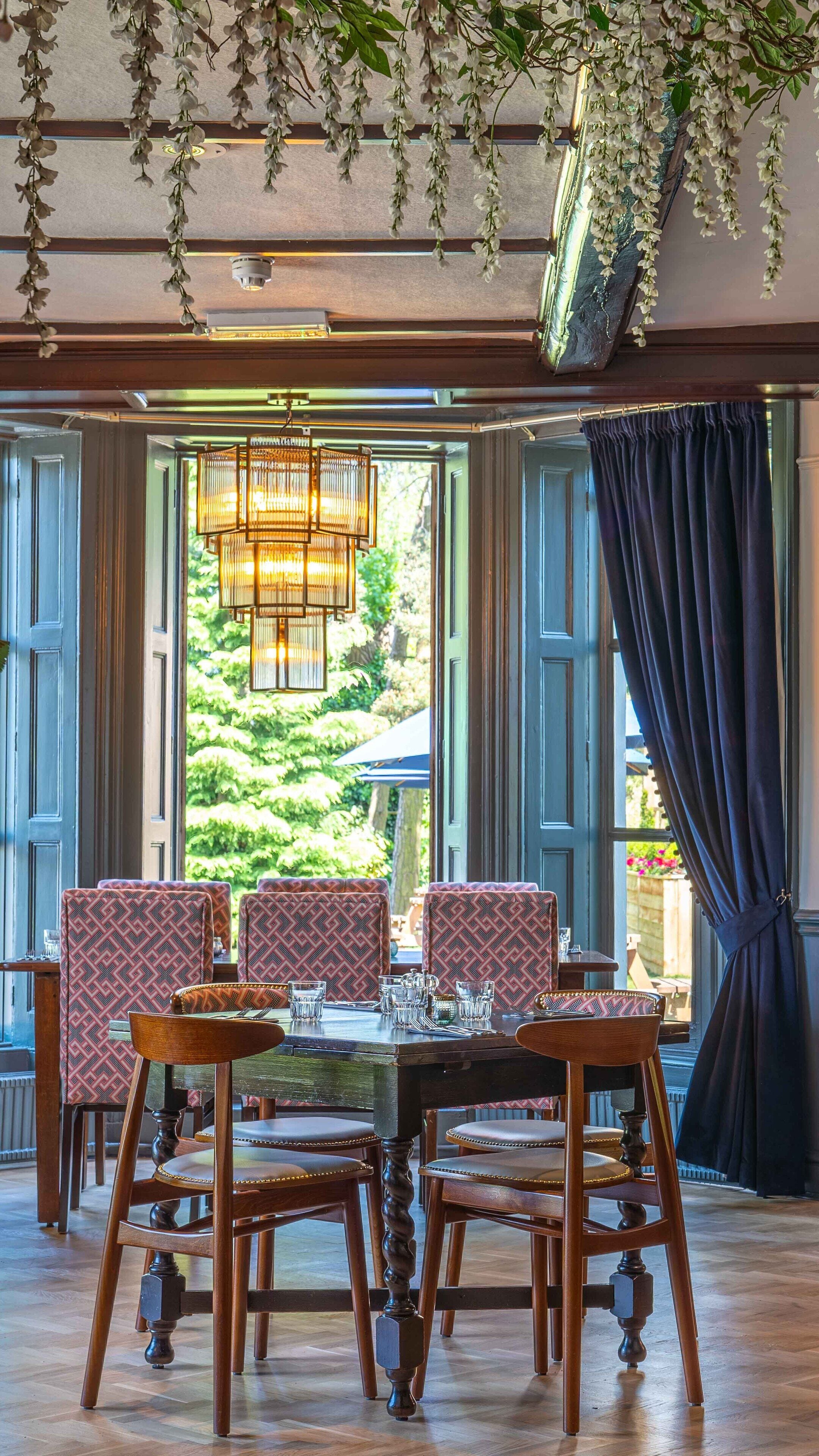 A dining area with a table set for four, surrounded by patterned chairs, a hanging light fixture, and a view of greenery outside.