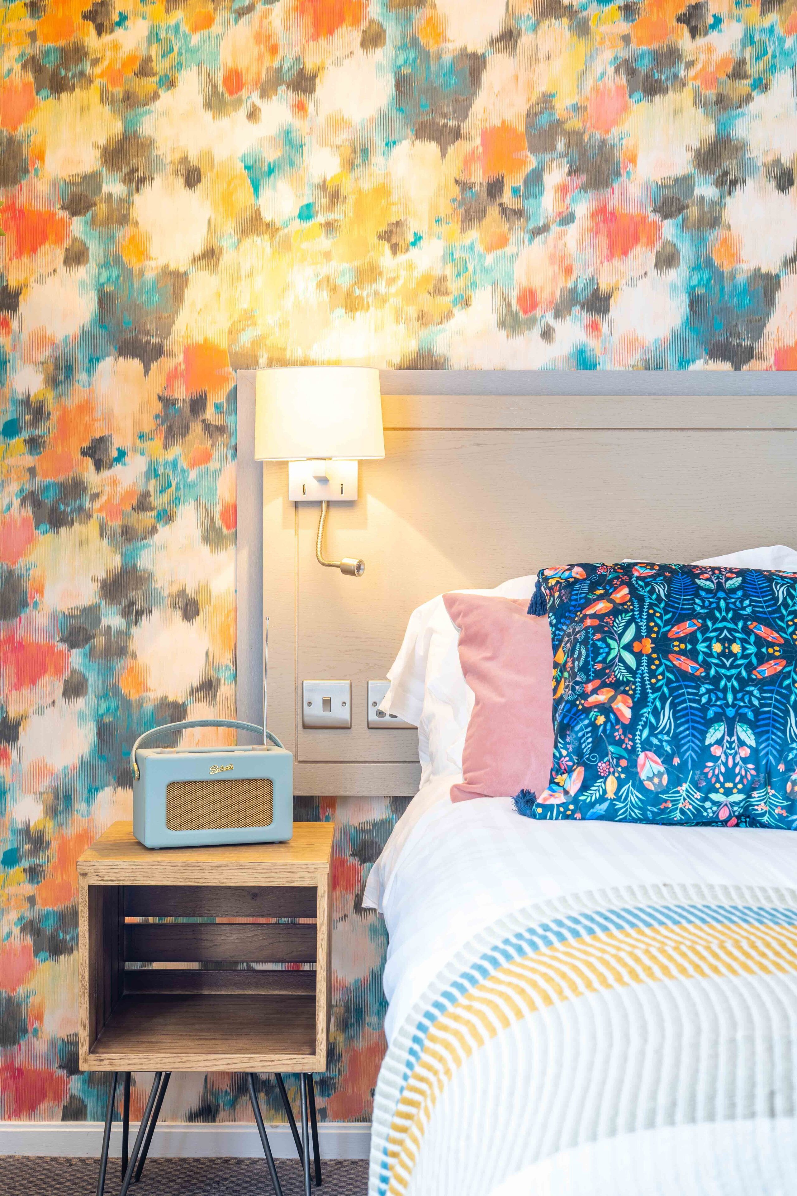 A hotel room with a colorful abstract wall mural, a bedside lamp, a radio, and a bed with a patterned pillow.