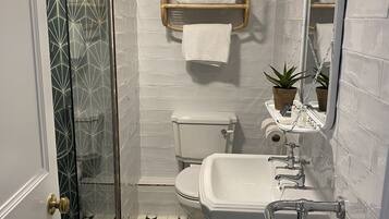 Un baño con inodoro, lavamanos y ducha. Hay un perchero con toallas y un pequeño estante con artículos de aseo.