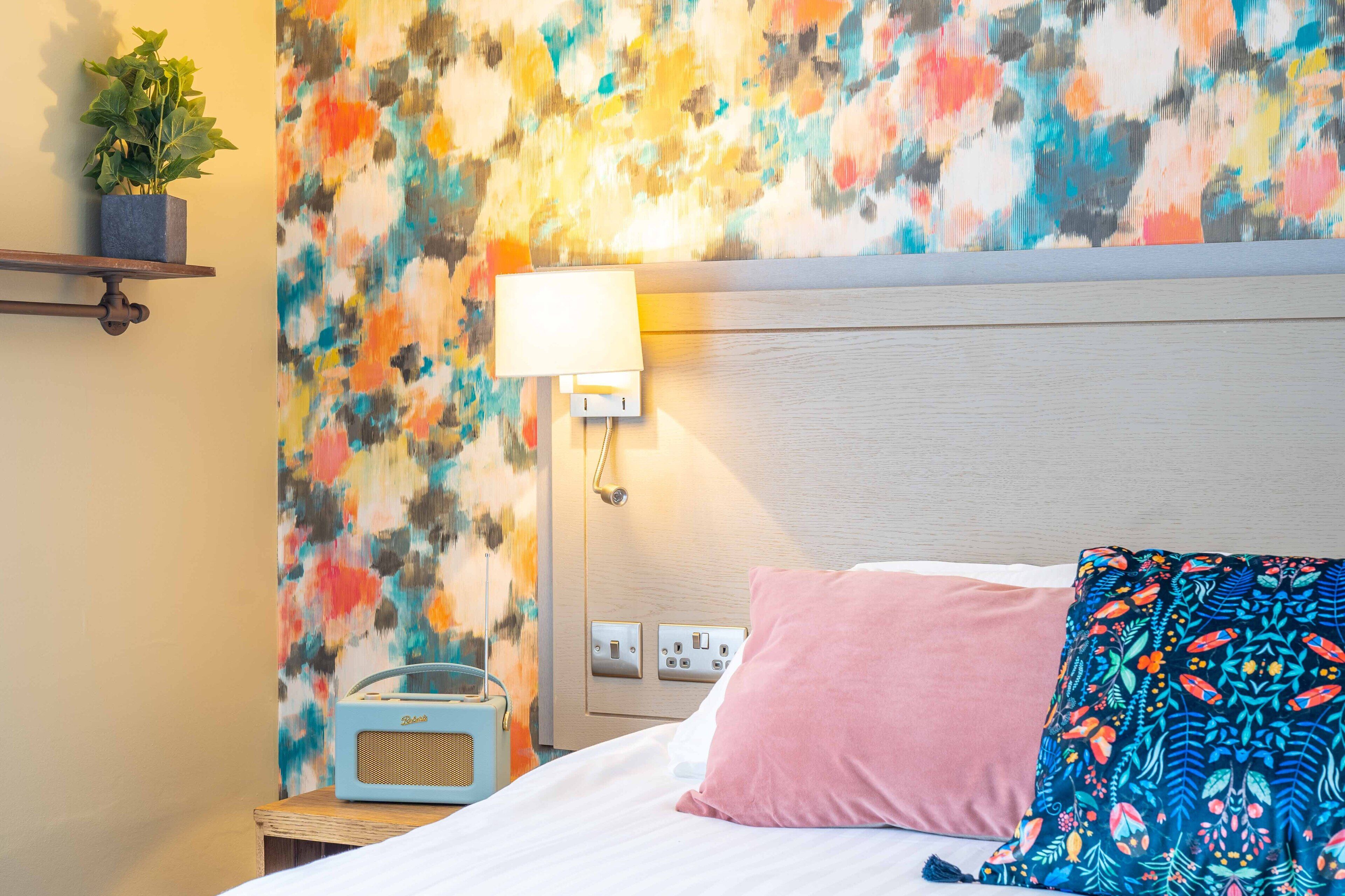 A hotel room with a bed, a bedside table with a lamp, a radio, and a colorful abstract wall.