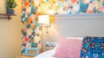 A hotel room with a bed, a bedside table with a lamp, a radio, and a colorful abstract wall.
