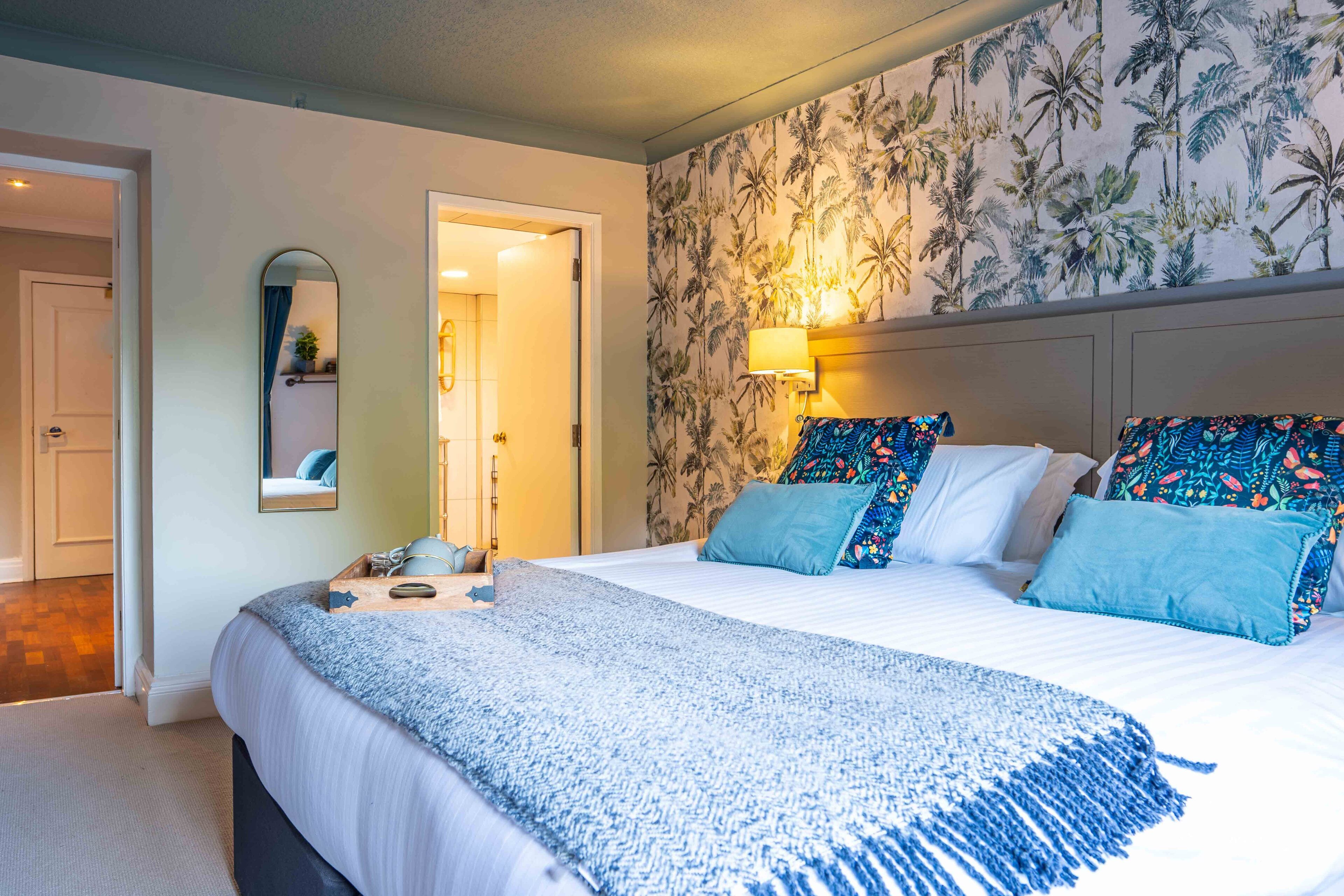 Um quarto com uma cama, um espelho, uma parede com papel de parede tropical e uma mesa de cabeceira com uma luminária.