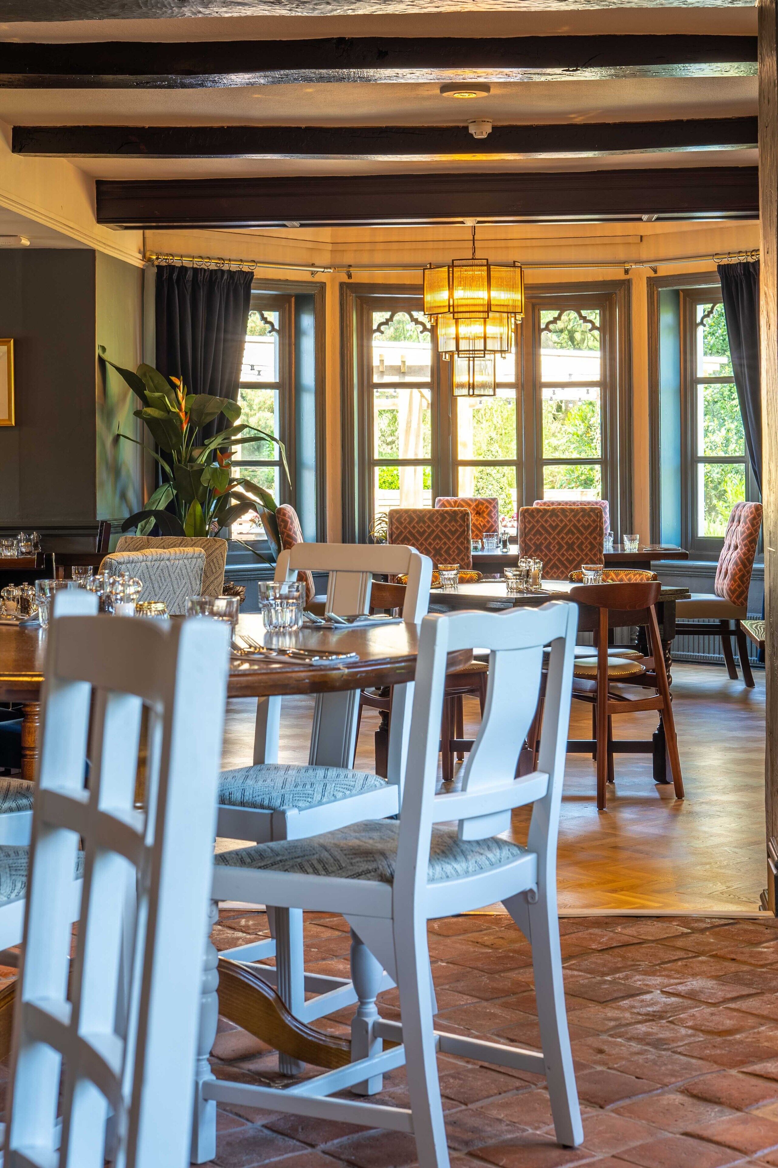A dining area with wooden tables and chairs, large windows with curtains, and a hanging light fixture.