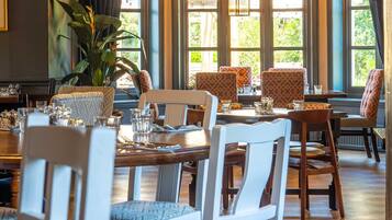 A dining area with wooden tables and chairs, large windows with curtains, and a hanging light fixture.