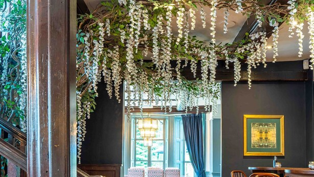 Un intérieur au plancher de bois, agrémenté de décorations florales suspendues, un espace repas avec des tables et des chaises, et un escalier avec des rampes en bois.