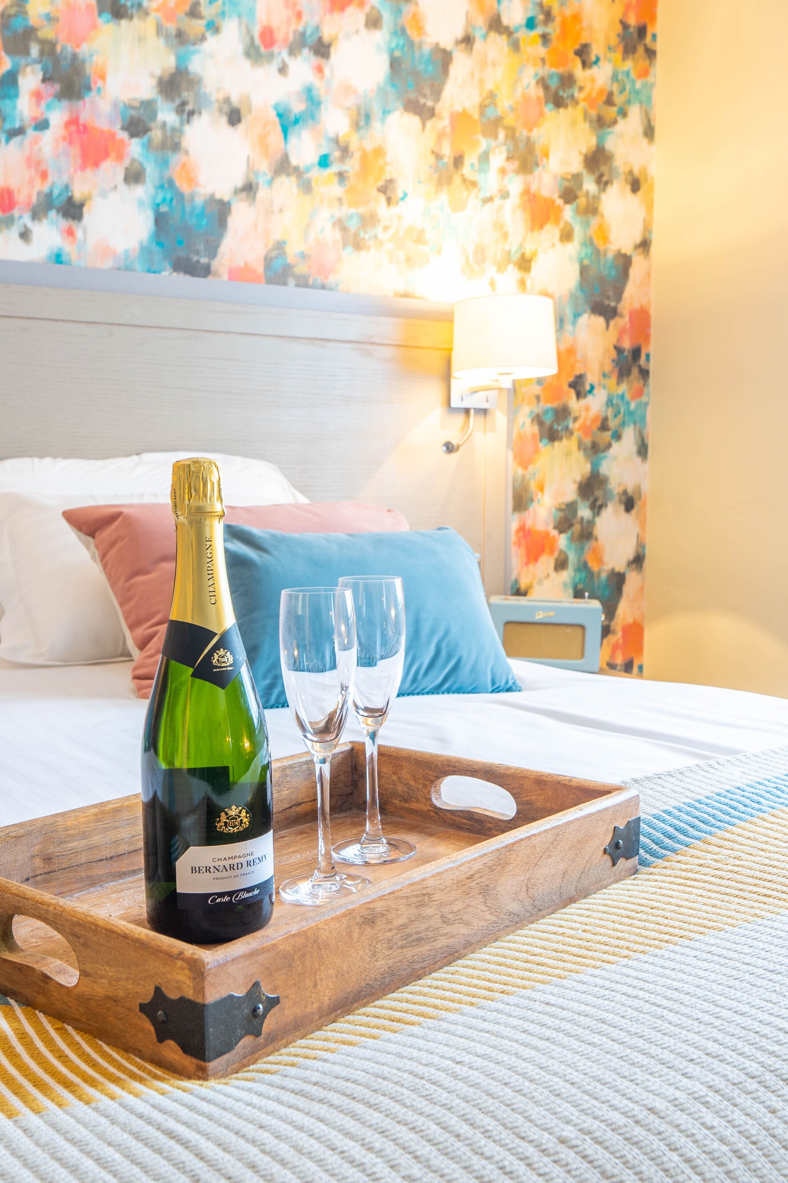 A bed with a wooden tray holding a bottle of champagne and two glasses, set against a colorful floral wall and a nightstand with a lamp.