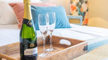 A bed with a wooden tray holding a bottle of champagne and two glasses, set against a colorful floral wall and a nightstand with a lamp.