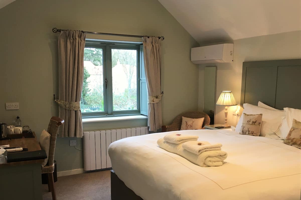 A hotel room with a large bed, a desk, a chair, a window with curtains, an air conditioning unit, and a lamp.