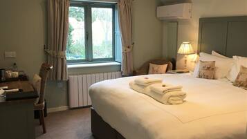 A hotel room with a large bed, a desk, a chair, a window with curtains, an air conditioning unit, and a lamp.