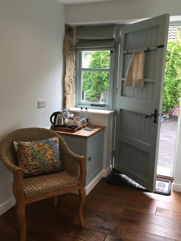 Habitación de esquina con una silla de madera, una mesita con utensilios de cocina, una ventana con vistas a zonas verdes y una puerta que da al exterior.
