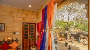 A room with a wooden wall, a sofa with cushions, a window with curtains, and a view of an outdoor area with trees and a person walking. - Boutique Helsinki (Jaisalmer)