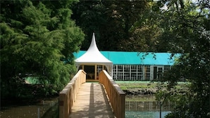 Een houten brug leidt naar een wit paviljoen met een puntige kap, omringd door groen en een waterpartij.