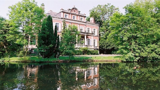 Een groot, historisch gebouw met een symmetrische gevel, omgeven door weelderige begroeiing en een rustig waterlichaam dat de structuur weerspiegelt.