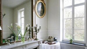 Een kamer met een open haard, een grote spiegel, een raam met uitzicht op bomen, en een tafel met boeken en een bloevas.