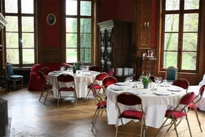 Ein Esszimmer mit runden Tischen für vier Personen, roten gepolsterten Stühlen und großen Fenstern mit Blick auf Grünflächen.