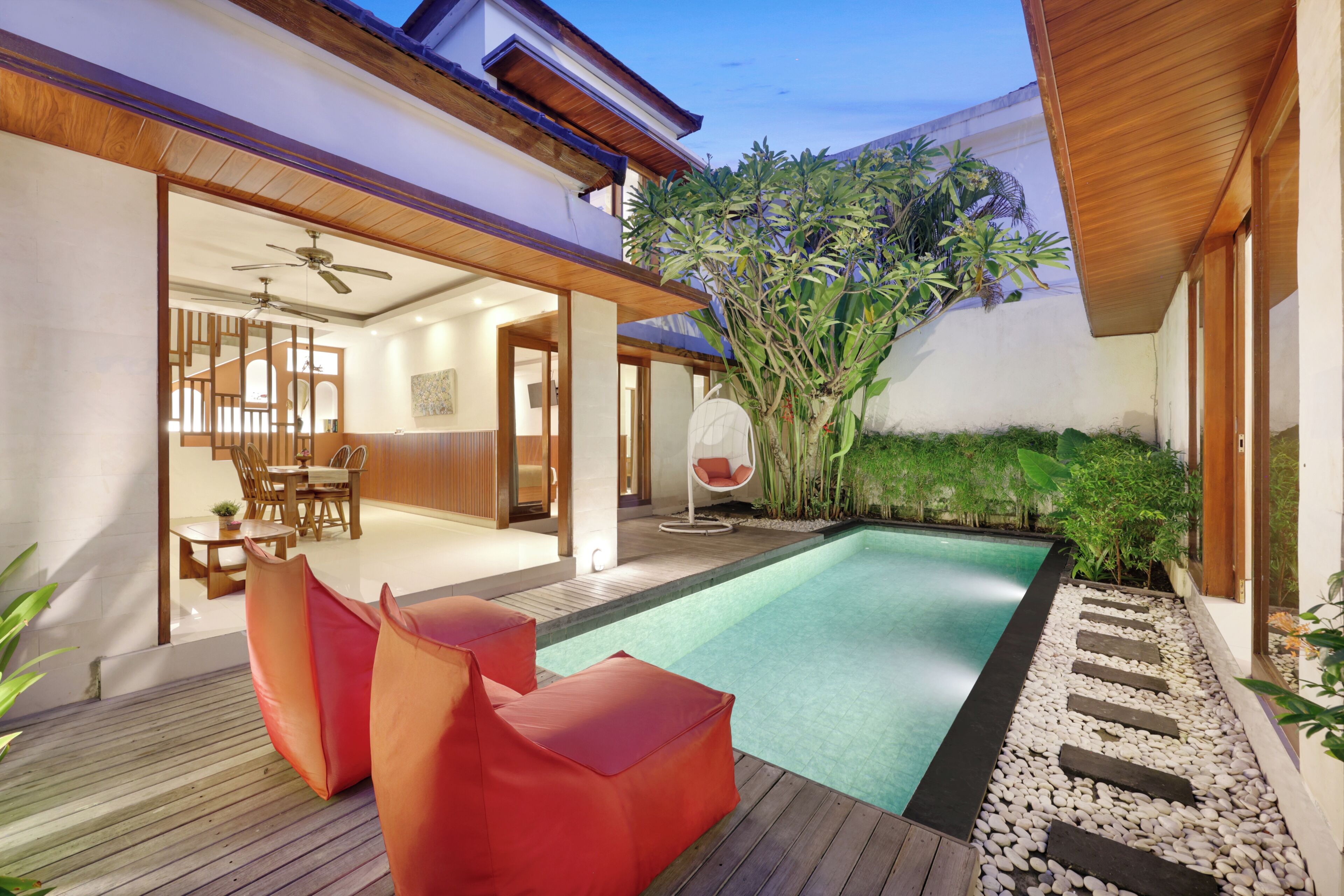 A modern outdoor pool area with a wooden deck, red seating, and a dining area visible through an open doorway.