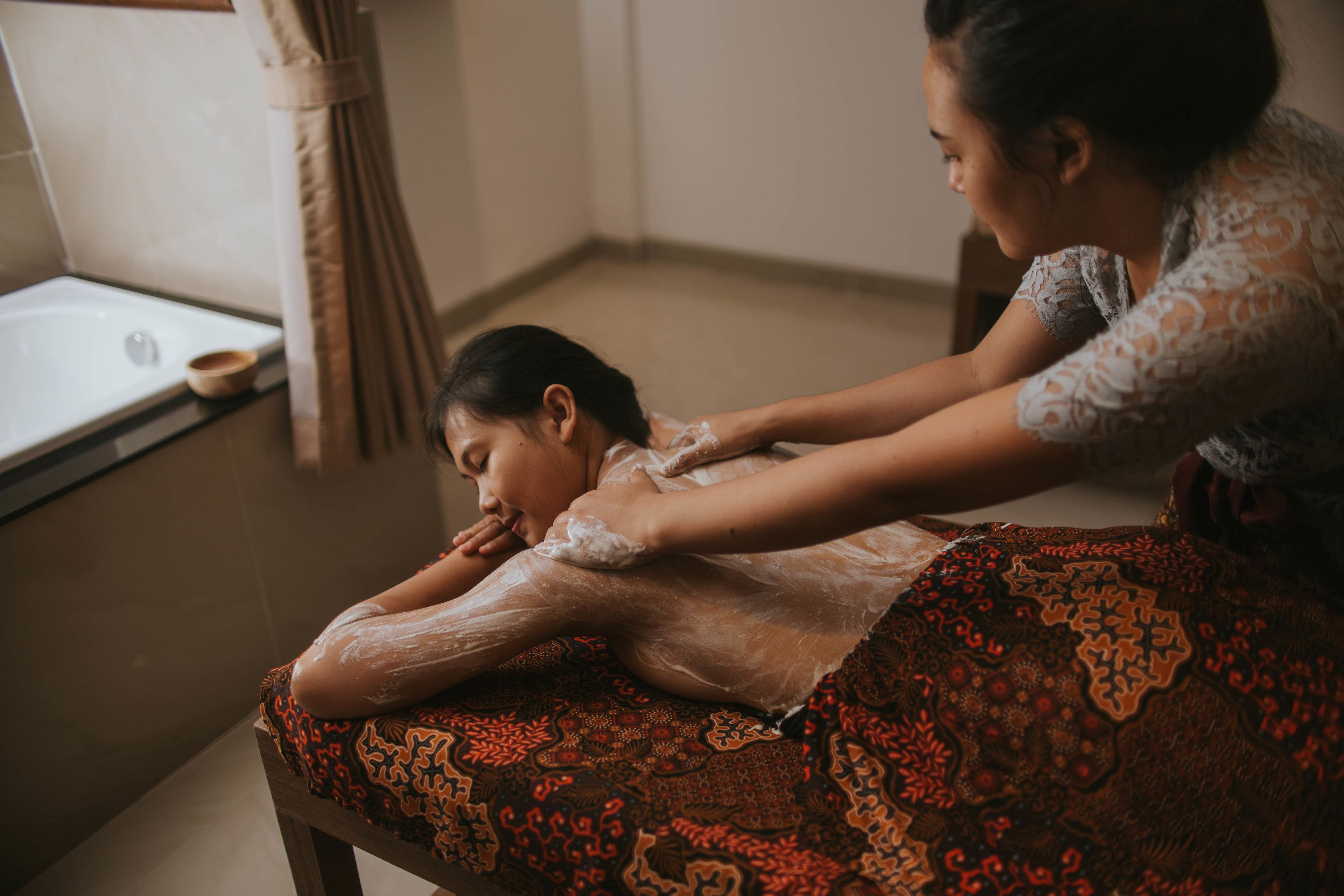 A person receiving a massage in a room with a bathtub and a window.