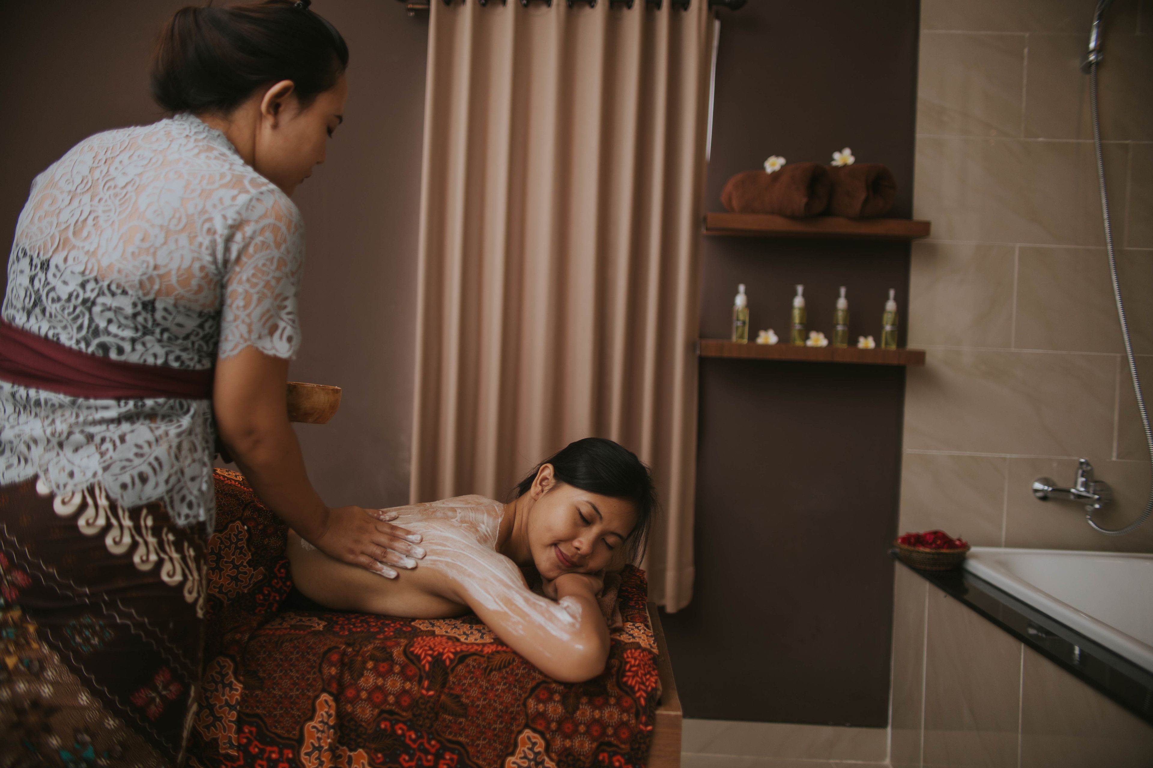 A person receiving a massage in a room with a bathtub, shelves with toiletries, and a curtain.