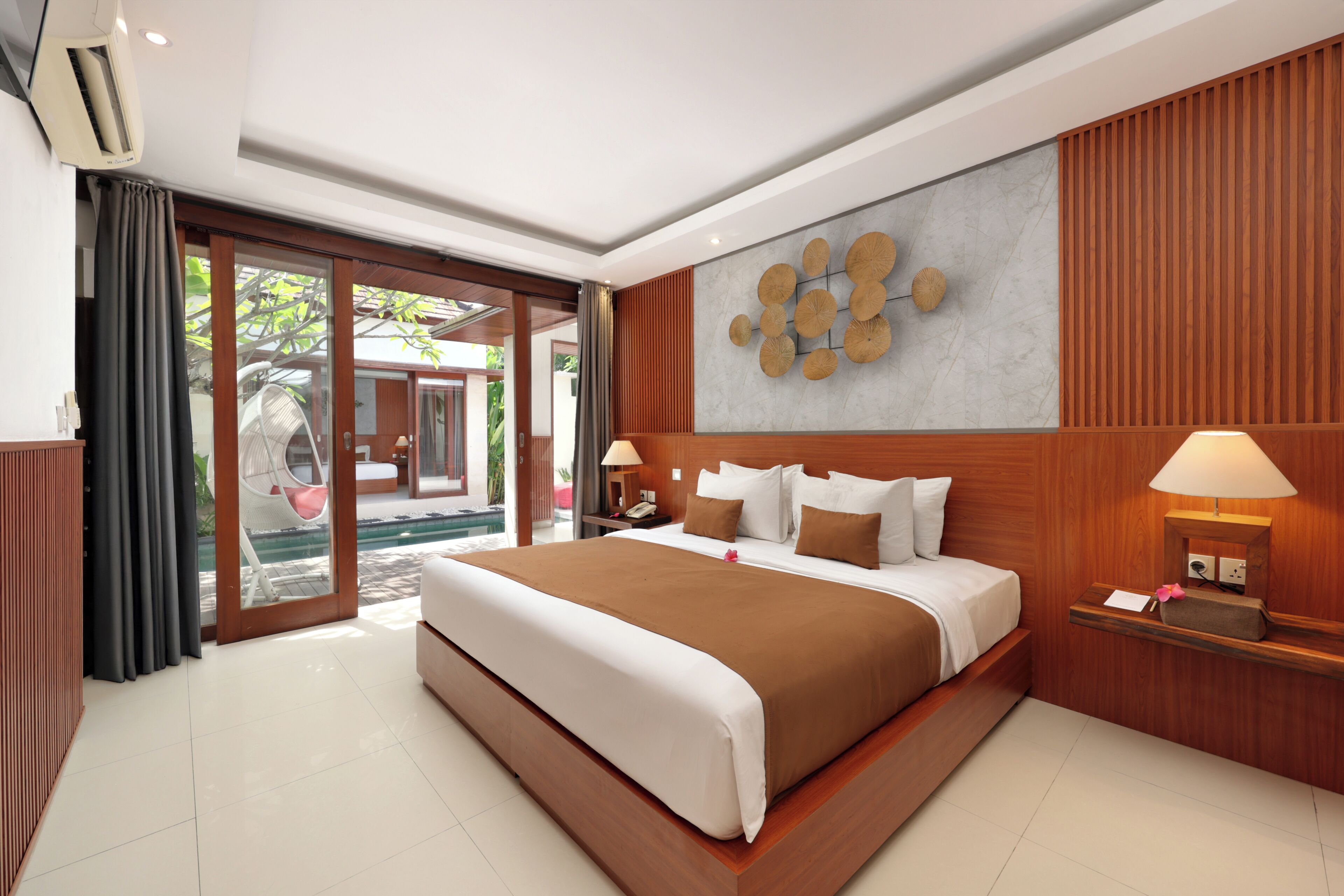 A modern hotel room with a large bed, a wooden headboard, a wall-mounted clock, and a view of a pool through a glass door.
