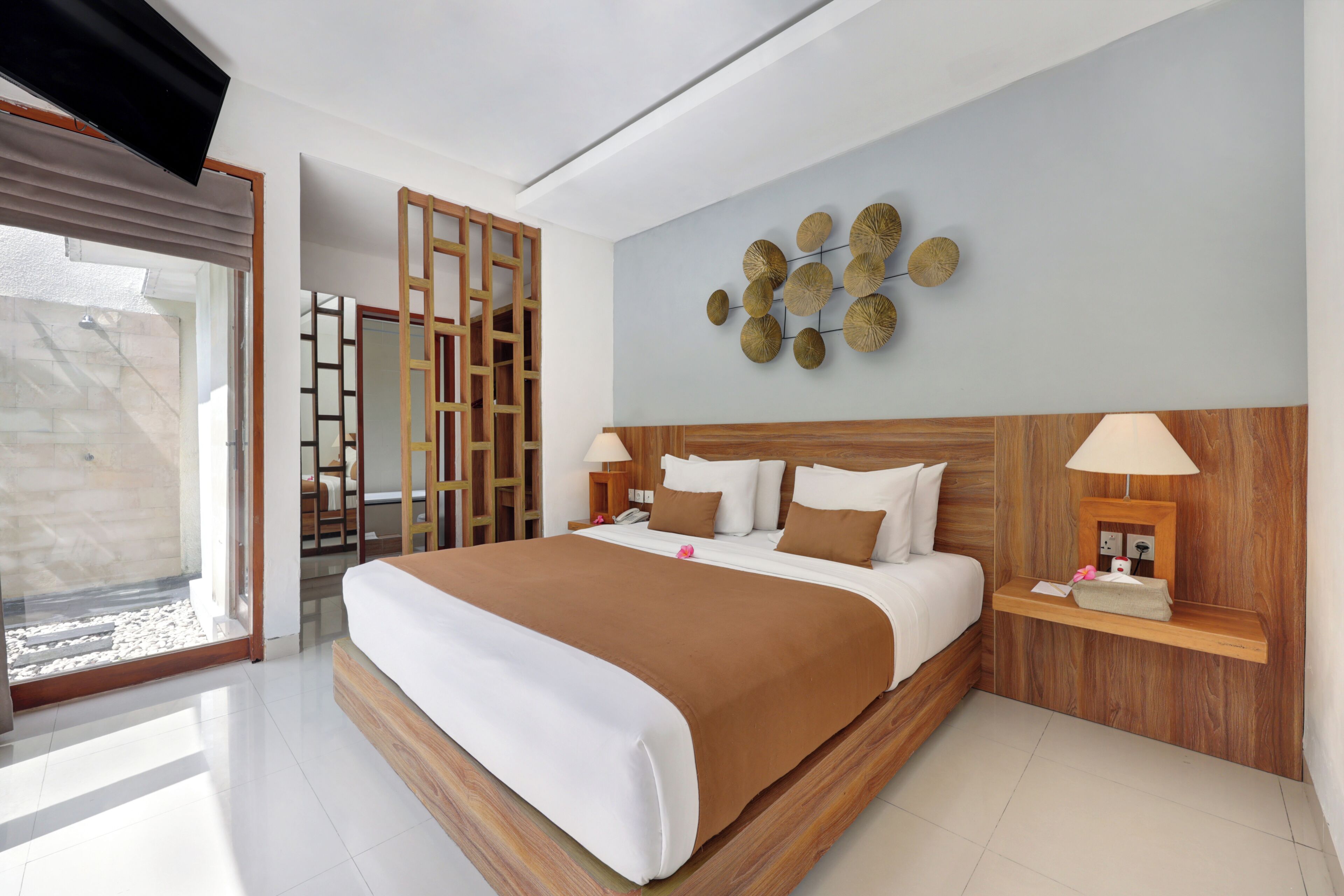 A modern hotel room with a large bed, wooden headboard, a wall-mounted decorative piece, and a bedside table with a lamp.