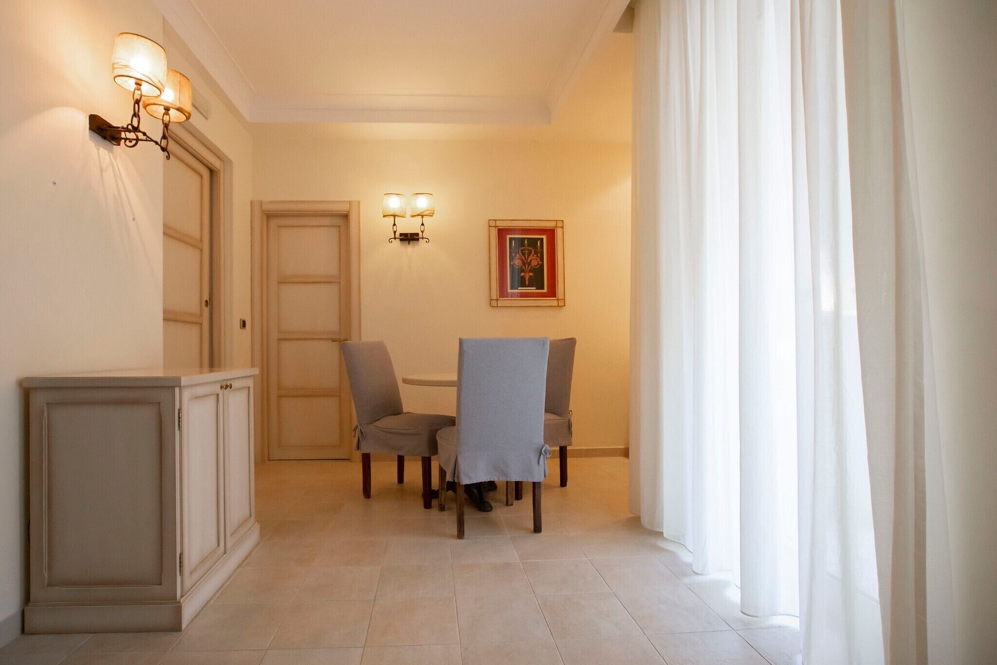 A hallway with a dining area featuring a table and chairs, a cabinet, wall-mounted lights, and a framed picture on the wall.