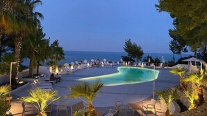 A pool area with lounge chairs and umbrellas, surrounded by palm trees and overlooking the sea. - Hotel Baia Dei Faraglioni (Mattinata)