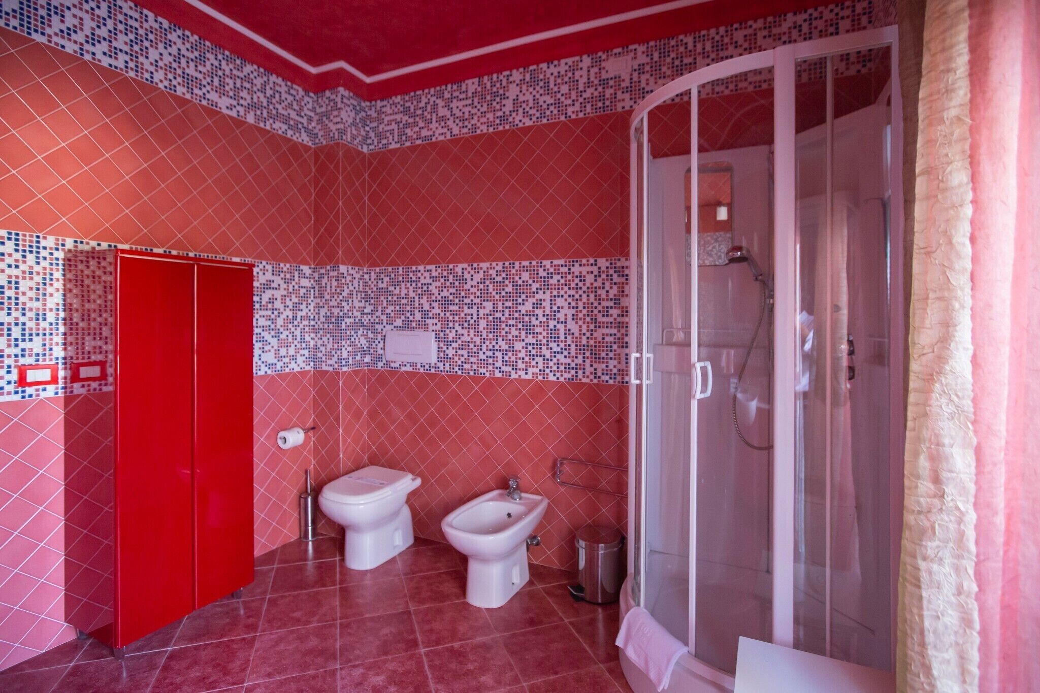 A bathroom with a red door, two bidets, a shower, and a tiled wall.