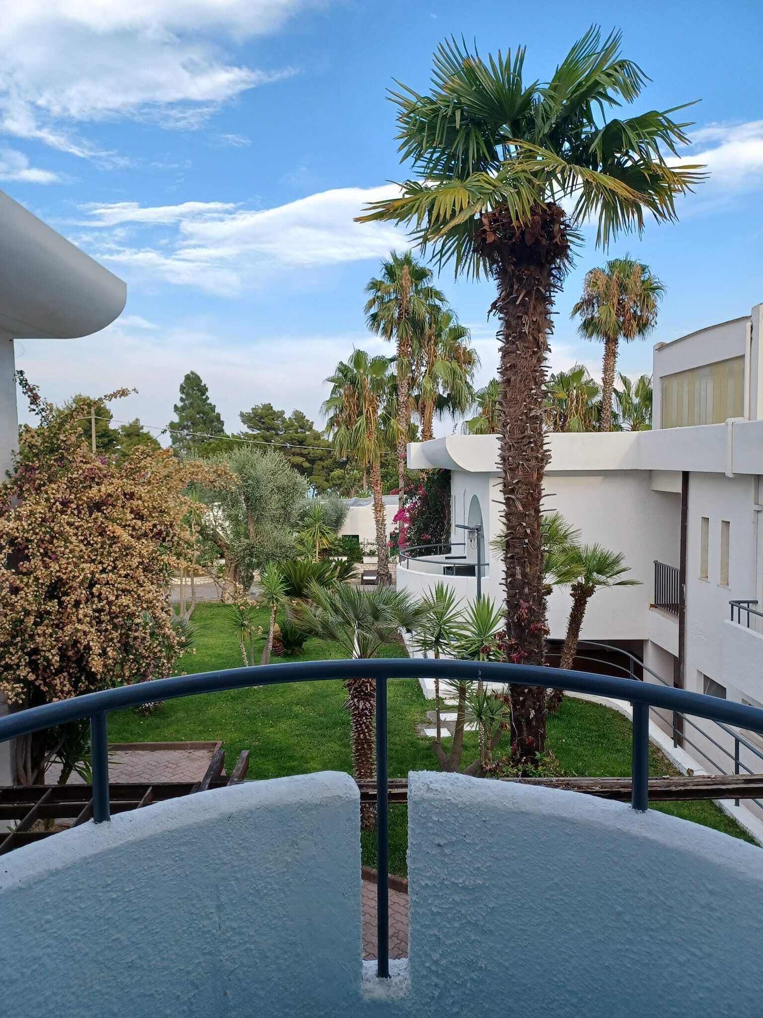Un balcon donnant sur un jardin aménagé, des palmiers et un bâtiment moderne.