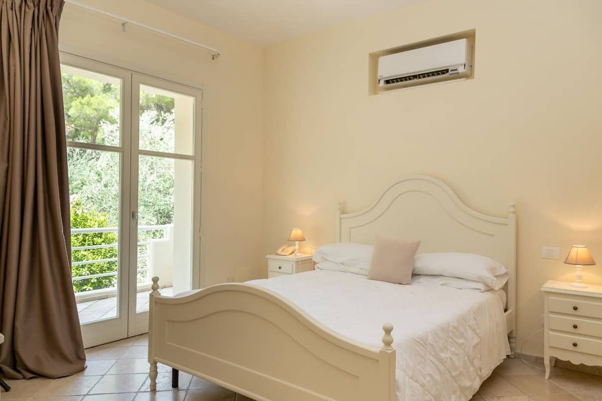 A bedroom with a bed, bedside tables, an air conditioning unit, and a view through a glass door.