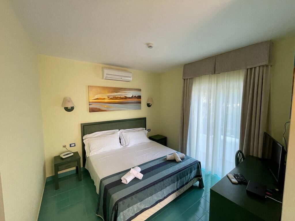 A hotel room with a bed, a desk with a computer, a TV, and a painting on the wall.