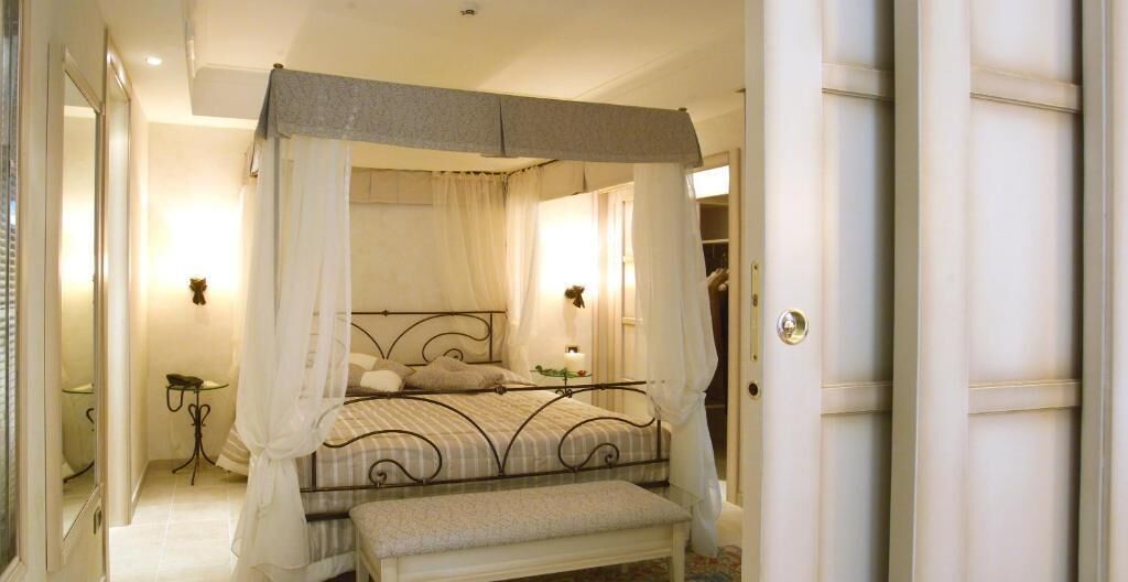 Una cama con dosel y cuatro columnas, un banco al pie, una mesita de noche y un espejo en la pared.