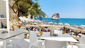 A beachside café with outdoor seating overlooking the sea, featuring white tables and chairs, a wooden deck, and a view of the coastline with rocky cliffs and a prominent rock formation. - Hotel Baia Dei Faraglioni (Mattinata)