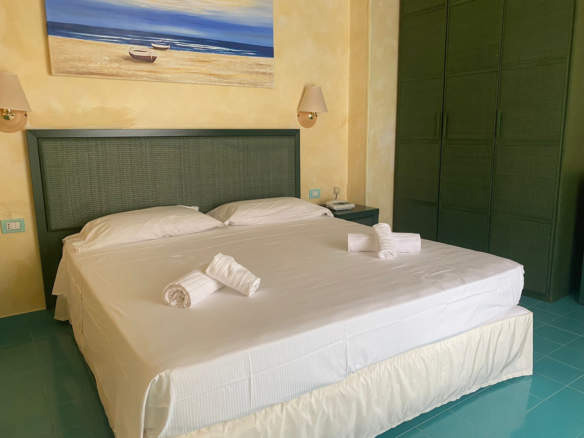 Un lit bien fait, avec du linge de lit et des serviettes blanches, un tableau représentant une scène de plage accroché au mur et une porte verte.