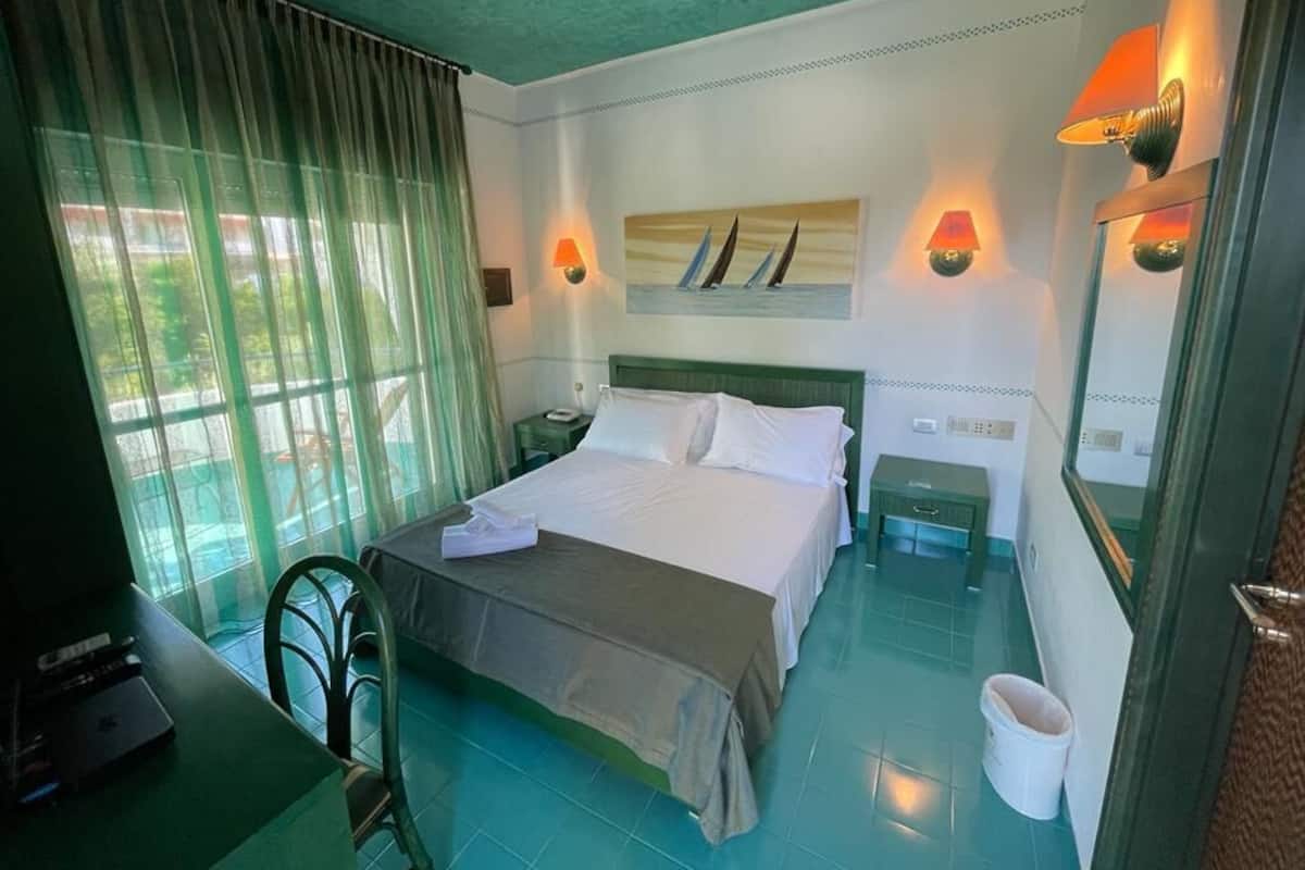A hotel room with a bed, a desk, a chair, a nightstand, a lamp, and a window with curtains.