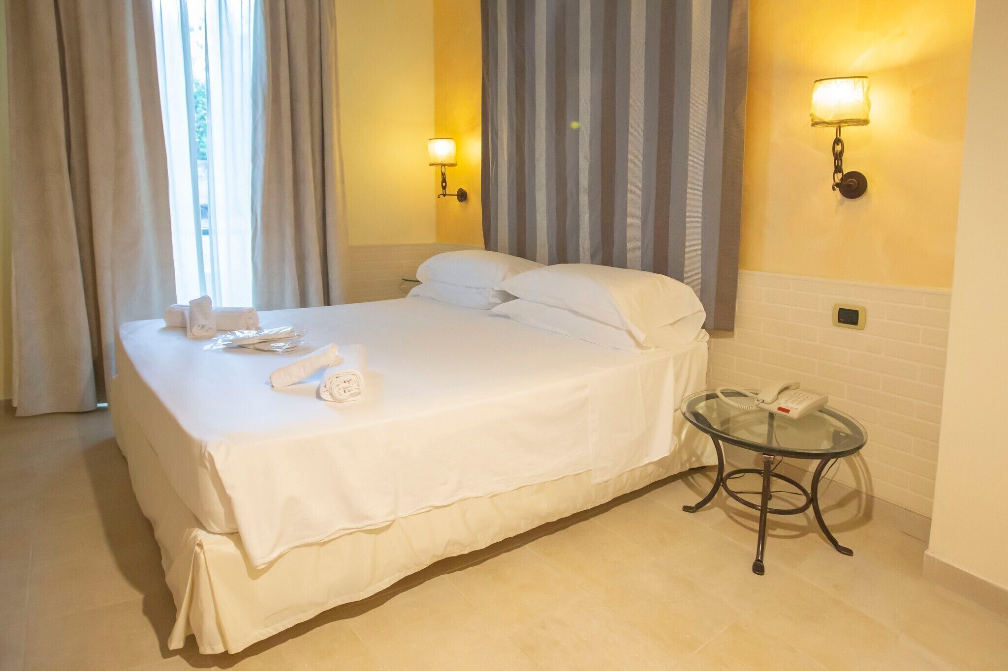 Una cama bien hecha con sábanas y almohadas blancas, una mesita de noche con teléfono, y una lámpara fijada a la pared.