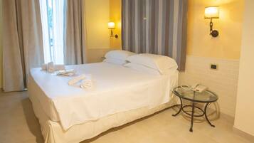 Una cama bien hecha con sábanas y almohadas blancas, una mesita de noche con teléfono, y una lámpara fijada a la pared.